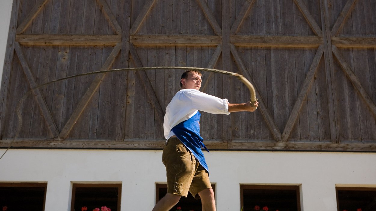 Die Goaslschneller und Schuahplattler Mann in traditioneller Kleidung schwingt eine Peitsche vor einem Holzgebäude