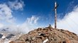Hotel a Santa Valburga in Val d’Ultimo Croce sulla vetta rocciosa della montagna sotto cielo blu e nuvole