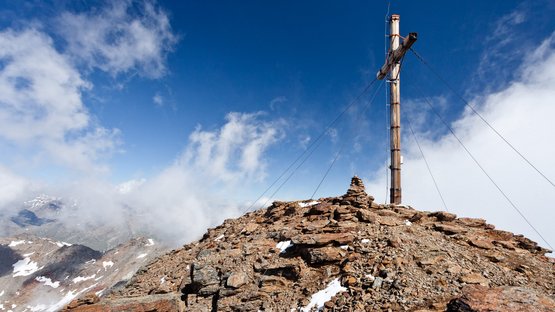 Hotel a Santa Valburga in Val d’Ultimo Croce sulla vetta rocciosa della montagna sotto cielo blu e nuvole