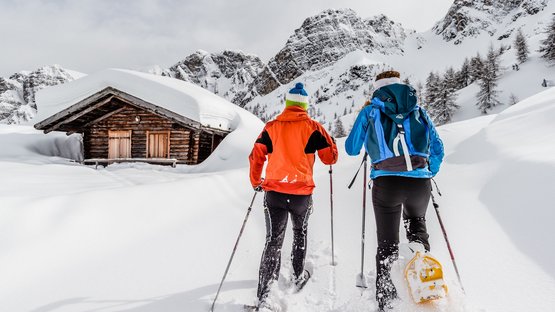 Schneeschuhwandern in Südtirol: Die Natur ruft! Zwei Personen beim Schneeschuhwandern vor einer schneebedeckten Hütte in den Bergen