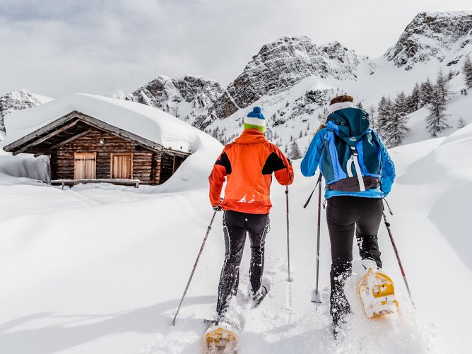 Schneeschuhwandern in Südtirol: Die Natur ruft! Zwei Personen beim Schneeschuhwandern vor einer schneebedeckten Hütte in den Bergen