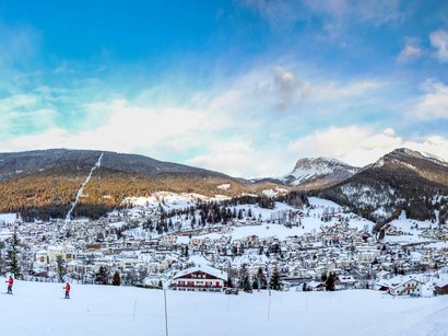 Hotels in Gröden in den Dolomiten! Winterpanorama einer verschneiten Stadt mit Bergen und Skifahrern im Vordergrund