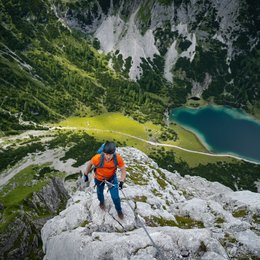 Pischadù-Klettersteig in Südtirol Kletterer in orange am Berg mit Blick auf See und grüne Landschaft