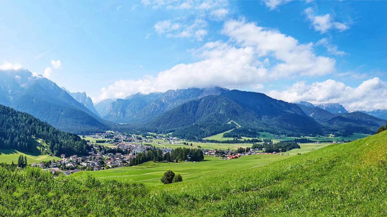 Prato Piazza: un gioiello della Val Pusteria Valle verde con villaggio davanti a montagne boschive sotto cielo azzurro