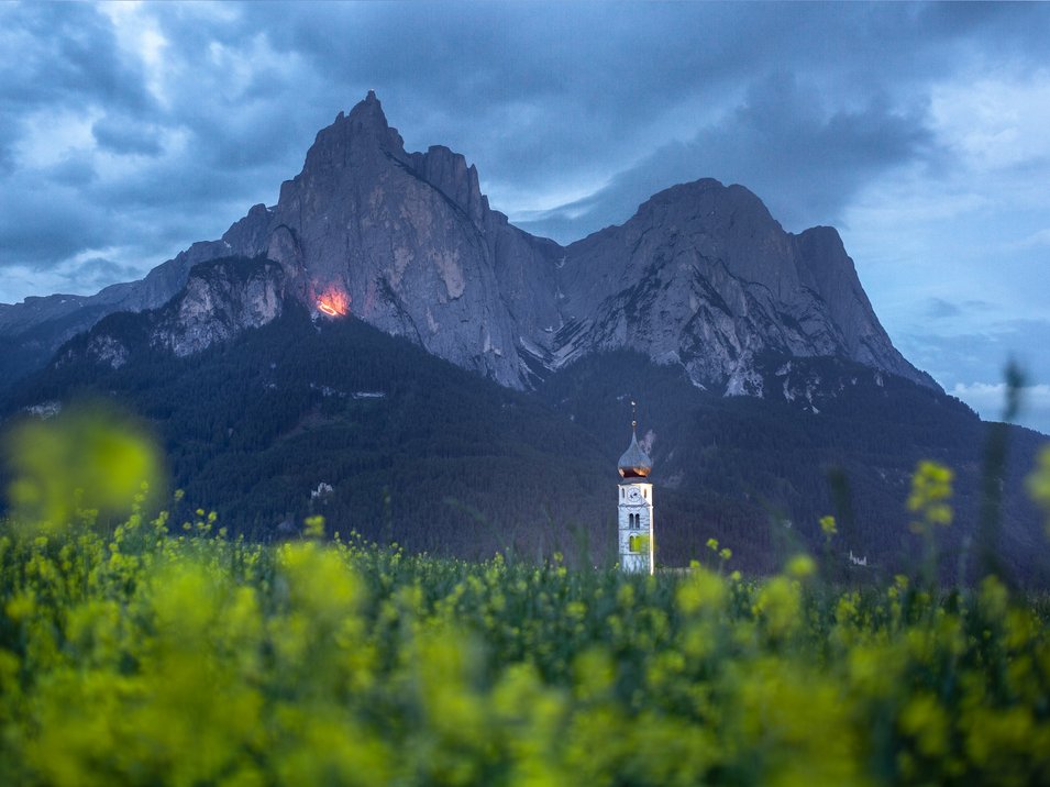 Herz-Jesu-Feuer in Südtirol Blumenfeld vor Berg und Kirchturm bei bewölktem Himmel am Abend
