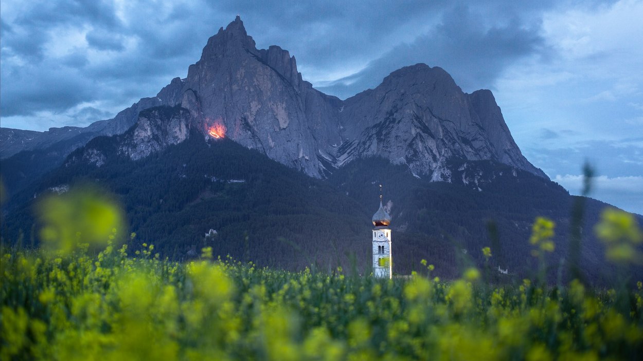 Herz-Jesu-Feuer in Südtirol Blumenfeld vor Berg und Kirchturm bei bewölktem Himmel am Abend