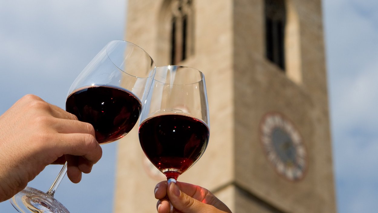 Weinhotels in Südtirol – Für Weinliebhaber Zwei Hände stoßen mit Rotweingläsern vor einer Turmuhr an