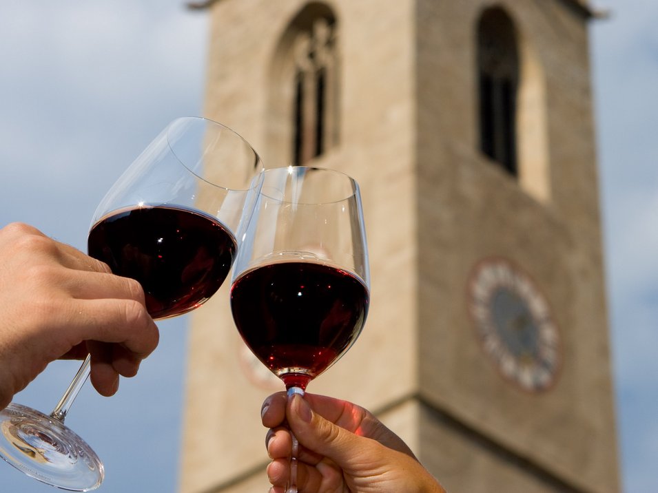 Hotel a Termeno sulla Strada del Vino Due mani brindano con calici di vino rosso davanti a una torre con orologio