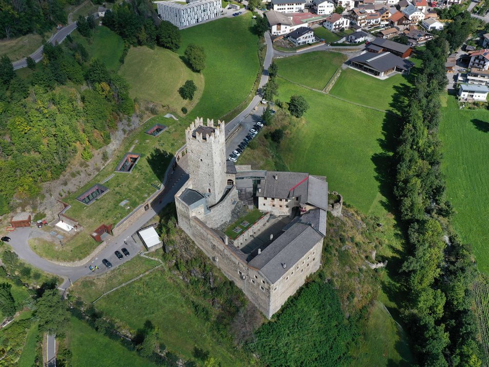 Die Fürstenburg bei Burgeis Luftaufnahme einer Burg auf einem Hügel umgeben von Grünflächen und Häusern