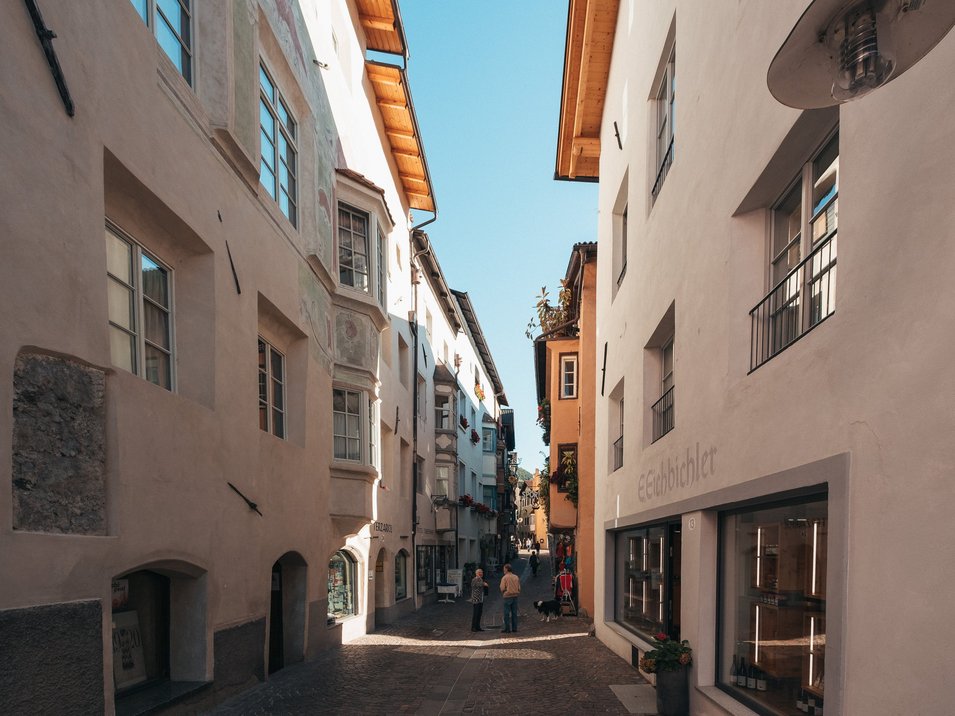 Hotels in Klausen: Ferien in Südtirol Menschen in einer engen, sonnigen Gasse mit historischen Gebäuden