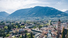 Hotels in St. Martin in Passeier: Ab in den Traumurlaub! Blick auf eine Stadt mit Kirche, Bergen im Hintergrund und bewölktem Himmel