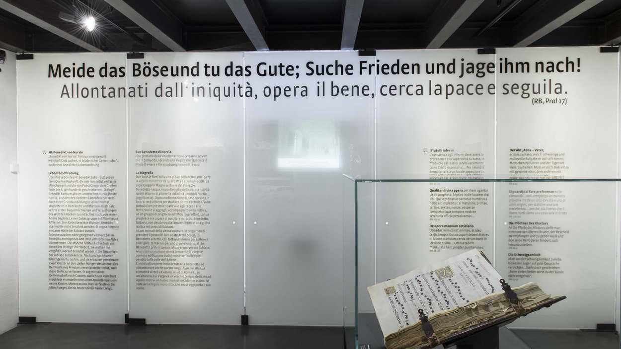 Das Benediktinerstift Marienberg Altes Buch in Vitrine vor Wand mit deutschem und italienischem Text zur Lebensbeschreibung