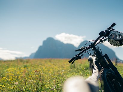 In mountain bike attraverso il Limojoch, il Tadegajoch e la Forcella da Cians passando per Fanes Mountain bike con casco in un prato fiorito davanti a montagne sotto cielo sereno