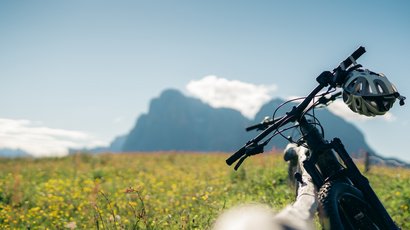 Circuito gourmet in Val Badia Mountain bike con casco in un prato fiorito davanti a montagne sotto cielo sereno
