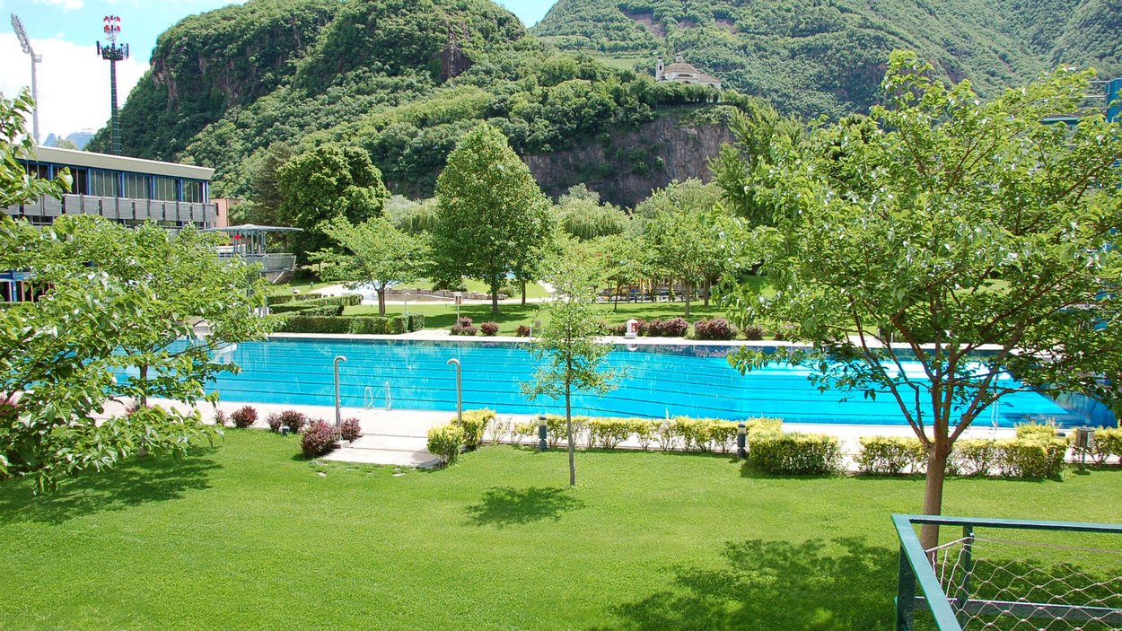Lido di Bolzano: punto di ritrovo per grandi e piccini Piscina e prato verde davanti a montagne boscose sotto cielo azzurro