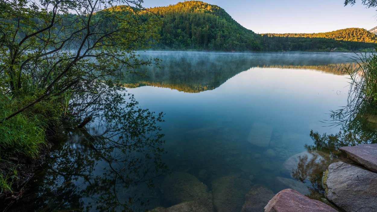 Montiggler Seen: Badespaß für Jung und Alt Berg und Bäume spiegeln sich in einem ruhigen See am Morgen