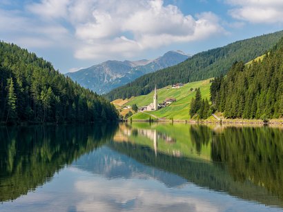 Al Lago di Favogna scoprirete una flora sorprendente Lago alpino con foresta e villaggio con chiesa nella valle sotto il cielo azzurro