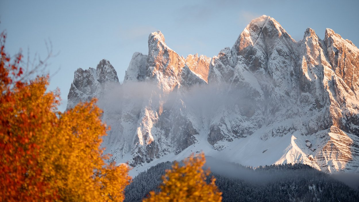 Divertirsi in Val di Funes Montagne innevate con alberi autunnali colorati e leggera nebbia