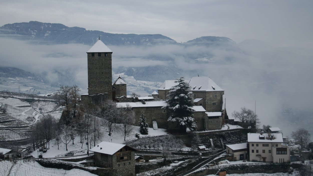Mercatino di Natale di Castel Tirolo Castello innevato su una collina con nebbia e montagne sullo sfondo