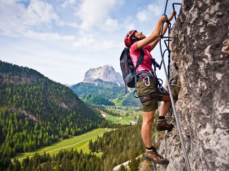 Hotels in Wolkenstein in Südtirol Frau klettert mit Helm und Ausrüstung an Felswand in den Bergen
