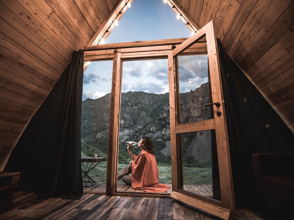 Glamping in Südtirol - Luxus & Natur Frau trinkt Wasser draußen sitzend vor offener Holzhüttentür mit Bergblick