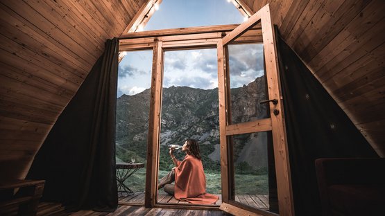 Chalets in Südtirol - Luxus & Ruhe Frau trinkt Wasser draußen sitzend vor offener Holzhüttentür mit Bergblick
