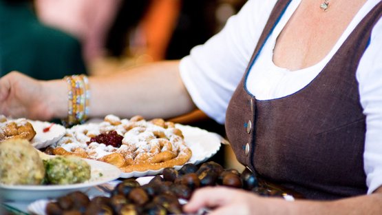 Bauernmärkte in Südtirol Frau trägt Teller mit verschiedenen traditionellen Speisen mit Puderzucker
