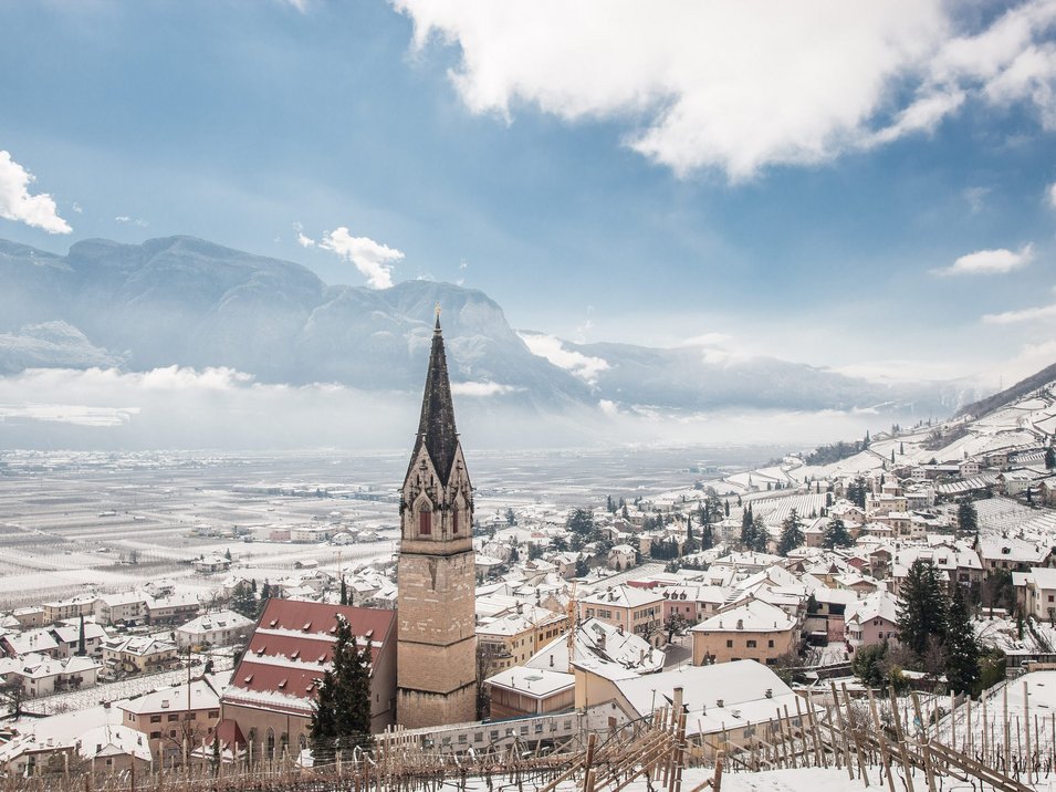 Hotel a Bolzano e dintorni Villaggio innevato con campanile davanti a montagne e cielo azzurro