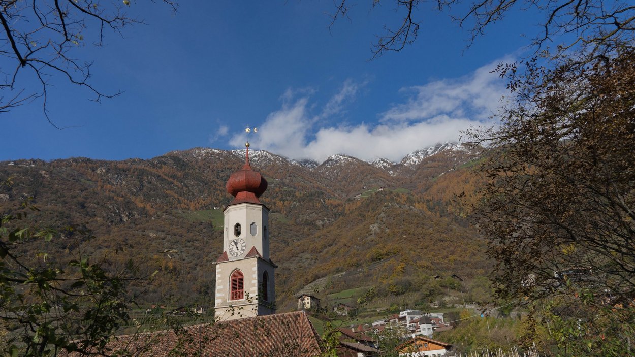Der Hoachwool-Klettersteig: gewaltig Kirchturm mit roter Kuppel vor herbstlichen Bergen und klarem blauen Himmel