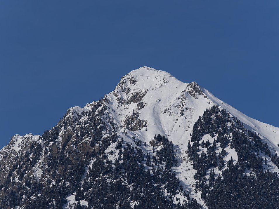 Wandern im Meraner Land Schneebedeckter Berg mit Nadelbäumen vor klarem blauem Himmel