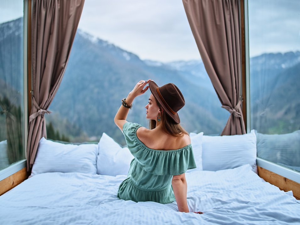 Boutiquehotels in Südtirol Frau mit Hut sitzt auf Bett vor Panoramafenstern mit Bergblick