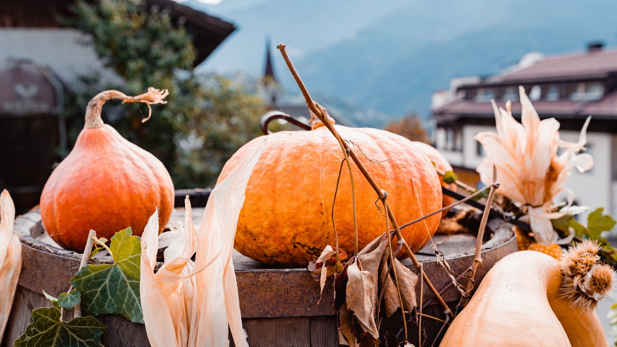 Roter Hahn - Schankbetriebe in Südtirol Herbstliche Kürbisse und getrocknete Pflanzen auf einem Holzbottich vor Bergkulisse