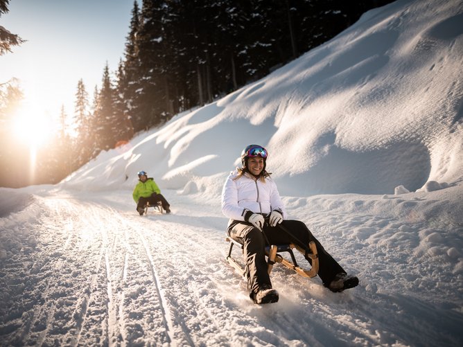 Winterurlaub in Südtirol: was für ein Genuss! Zwei Personen rodeln bei sonnigem Winterwetter auf verschneiter Piste im Wald