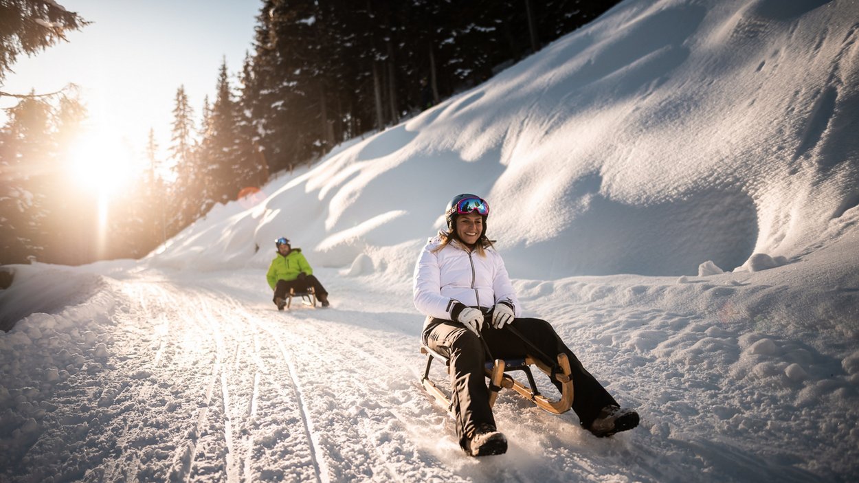 Die längste Rodelbahn Südtirols Zwei Personen rodeln bei sonnigem Winterwetter auf verschneiter Piste im Wald