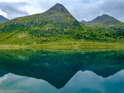 Hotels in St. Lorenzen: willkommen im Pustertal! Berg spiegelt sich im klaren See unter bewölktem Himmel