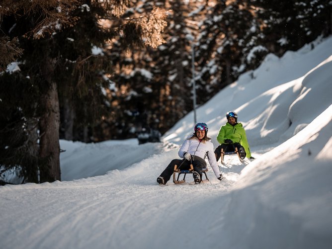 Slittino in Alto Adige: come far battere forte il cuore. Due persone fanno slittino felicemente su percorso innevato nella foresta