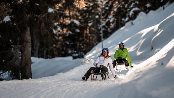 Rodeln in Südtirol: da schlägt das Herz schneller. Zwei Personen rodeln fröhlich auf verschneiter Strecke im Wald