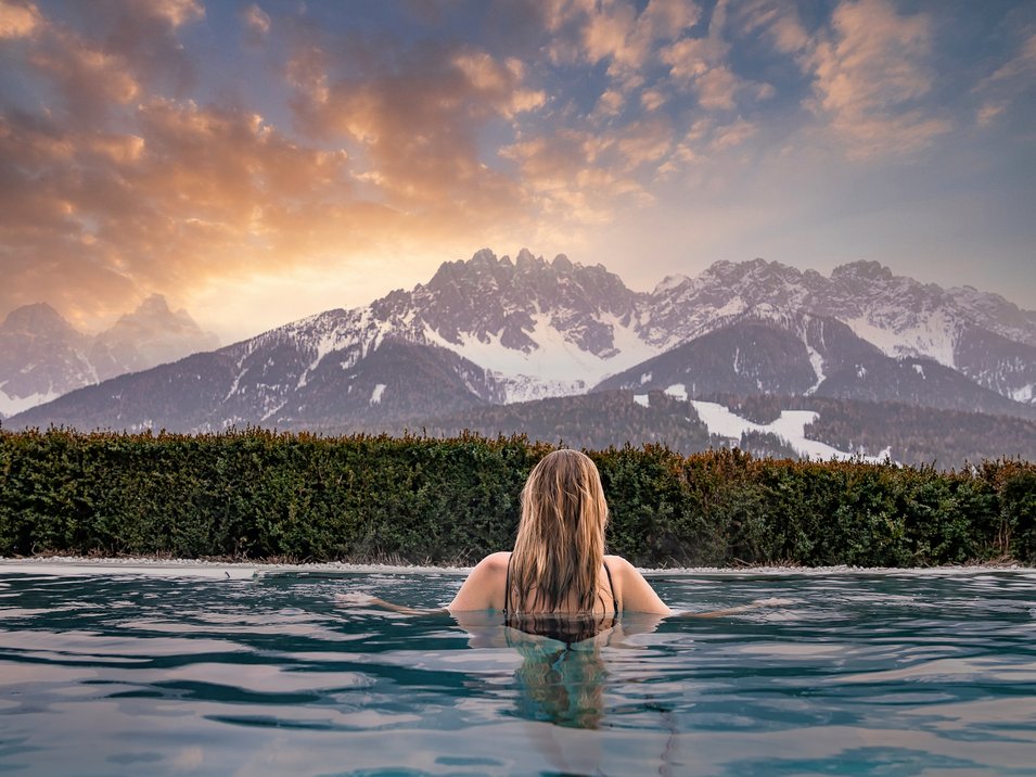 5-Sterne-Hotel in Südtirol gesucht? Gefunden! Frau schwimmt im Pool mit Blick auf schneebedeckte Berge bei Sonnenuntergang