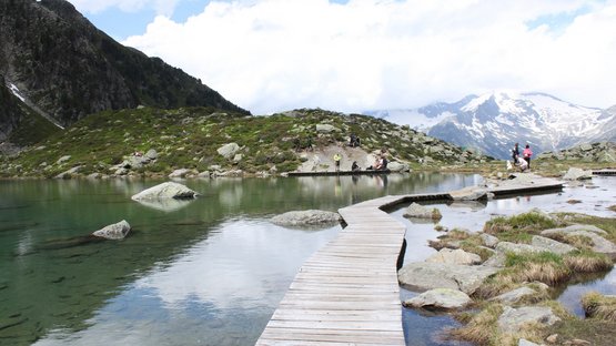 Hotel a Cadipietra in Alto Adige Passerella in legno su lago di montagna con escursionisti e montagne innevate sullo sfondo