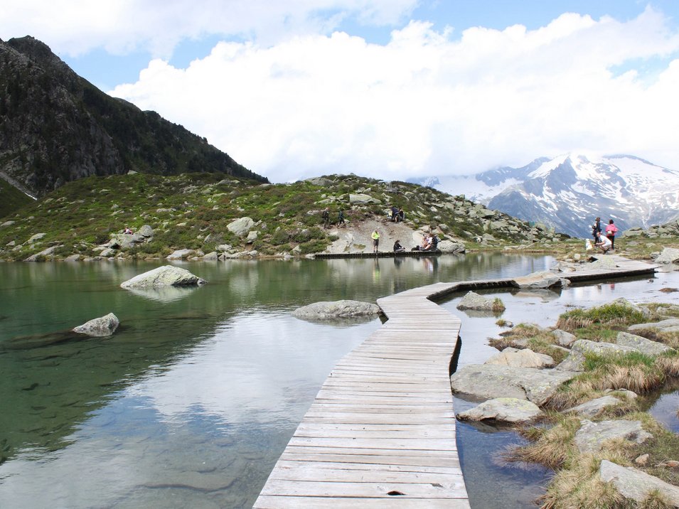 Hotel a Cadipietra in Alto Adige Passerella in legno su lago di montagna con escursionisti e montagne innevate sullo sfondo