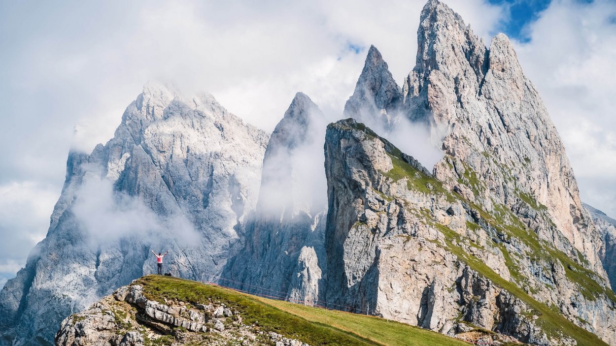 Sass Rigais – unterwegs in der Geislergruppe Wanderer auf grünem Hügel vor beeindruckenden Dolomitenfelsen und Nebel