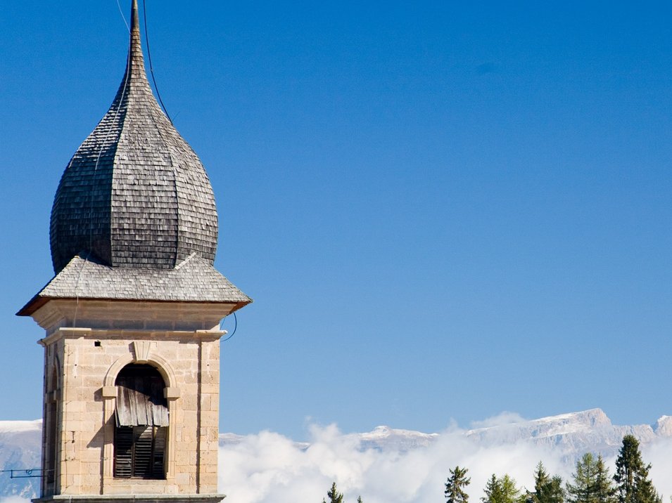 Hotels in Deutschnofen: Urlaub in den Dolomiten Alter Kirchturm vor bewaldeten Bergen und blauem Himmel in den Alpen