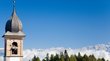 Hotel a Nova Ponente Vecchio campanile davanti a montagne boschive e cielo azzurro nelle Alpi