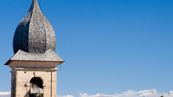 Hotels in Deutschnofen: Urlaub in den Dolomiten Alter Kirchturm vor bewaldeten Bergen und blauem Himmel in den Alpen