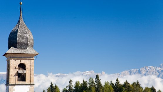 Hotel a Nova Ponente Vecchio campanile davanti a montagne boschive e cielo azzurro nelle Alpi