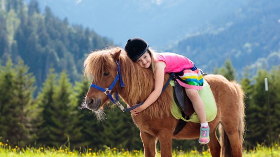 Südtiroler Familienhotels – Lust auf einen Tapetenwechsel? Mädchen in Helmkappe umarmt ein Pony im Bergwiesenfeld mit Wald