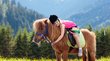 Family hotel in Alto Adige Bambina con casco abbraccia un pony in un prato di montagna con foresta