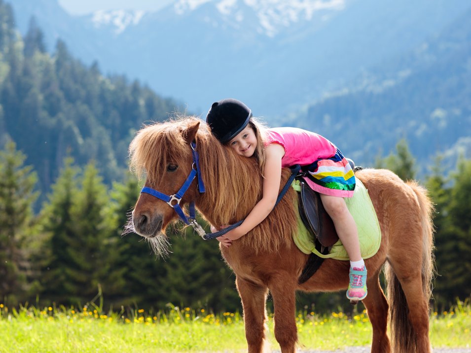 Family hotel in Alto Adige Bambina con casco abbraccia un pony in un prato di montagna con foresta