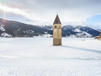 Langlaufen in Südtirol – in Sulden am Ortler Kirchturm ragt aus zugefrorenem See in verschneiter Berglandschaft