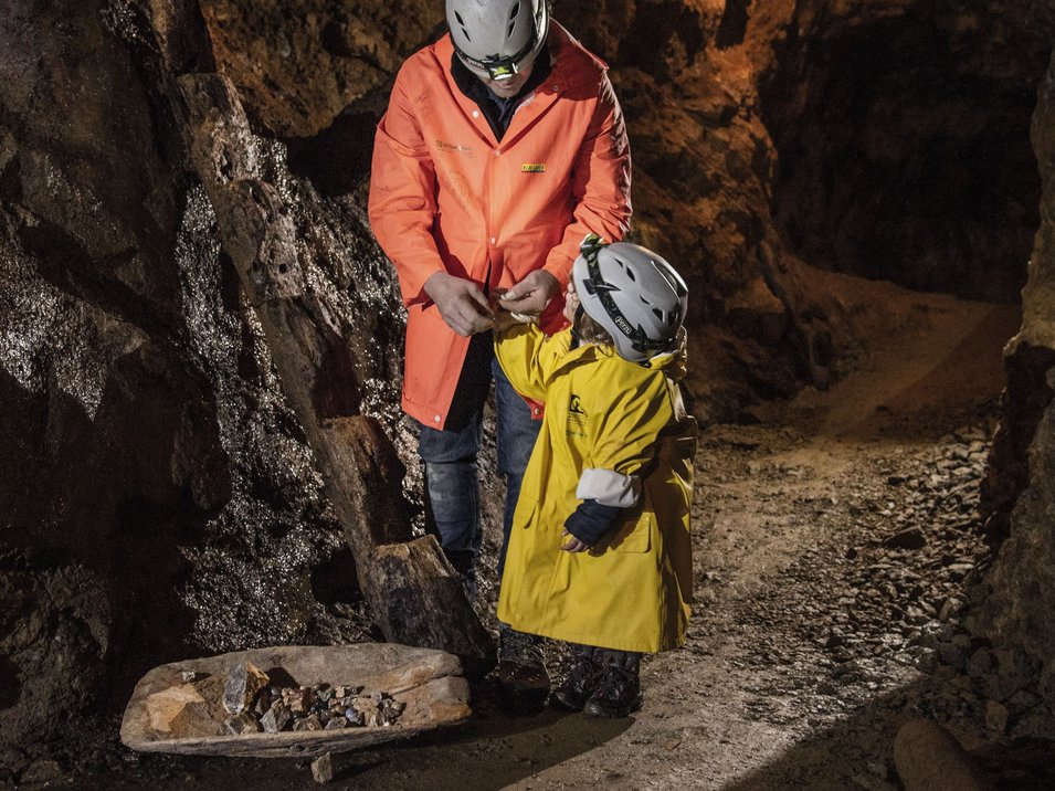 Bergwerk Villanders – ein Stück Geschichte Erwachsener und Kind in Helmen und Jacken in einer minenhöhle mit Erzstücken